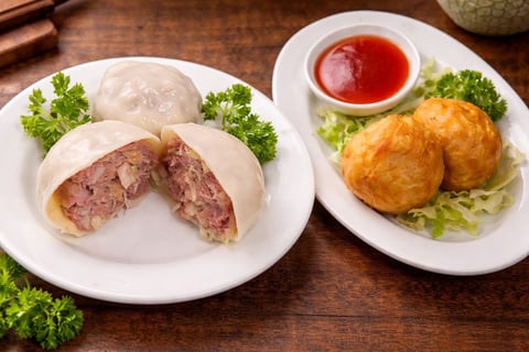 Two plates of Asian cuisine on a wooden table: steamed dumplings with pork filling on the left, and fried spring rolls with dipping sauce on the right, garnished with fresh parsley