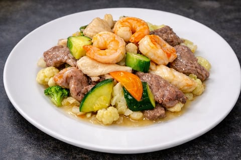Stir-fry dish on white plate with shrimp, beef, broccoli, cashews, and bell peppers on dark surface