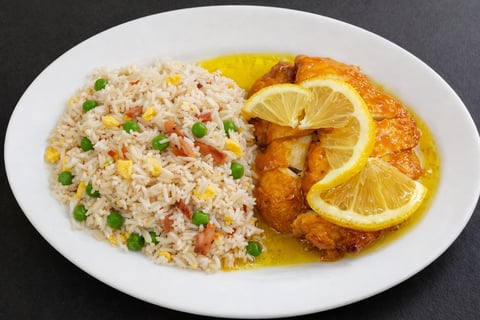 A white plate with golden-brown breaded chicken fillet topped with lemon slices beside a serving of fried rice with peas and carrots