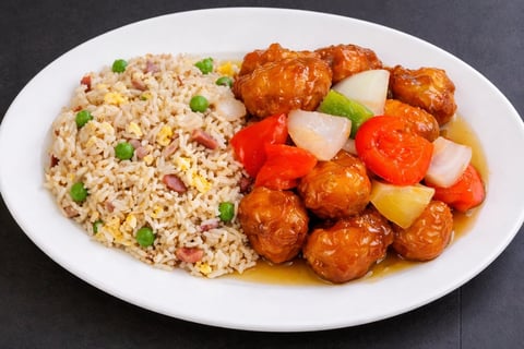 Plate of fried rice with peas and carrots beside sweet and sour meatballs with peppers and pineapple
